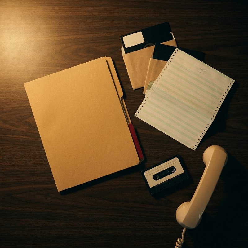 A plain manila folder on a laminate desk beside floppy disks, striped printer paper, a cassette tape and a beige telephone handset.